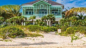 A turquoise two-story house with a balcony, surrounded by palm trees and a sandy beach.