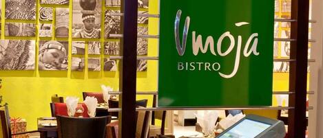 Interior de un restaurante con un letrero verde que dice 'Vmoja BISTRO', mesas con servilletas blancas y una pantalla digital con el menú.