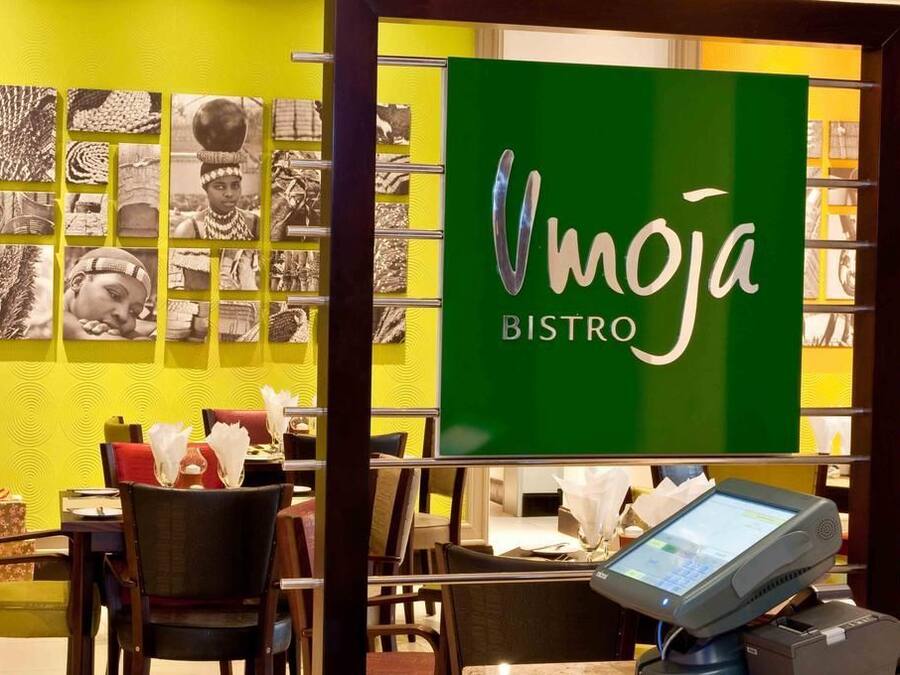 A restaurant interior with a green sign reading 'Vmoja BISTRO,' tables with white napkins, and a digital menu display.