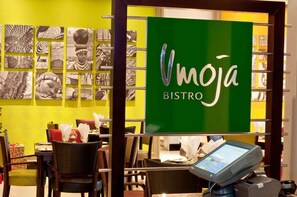 L’intérieur d’un restaurant, avec une enseigne verte portant l’inscription « Vmoja BISTRO », des tables avec des serviettes blanches et un menu affiché sur un écran numérique.
