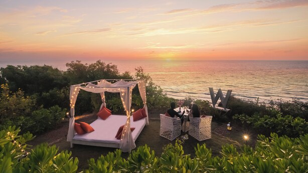 Una cama con dosel y almohadas, una mesa con sillas, y vista al mar al atardecer.
