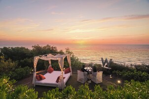 Una cama con dosel y almohadas, una mesa con sillas, y vista al mar al atardecer.
