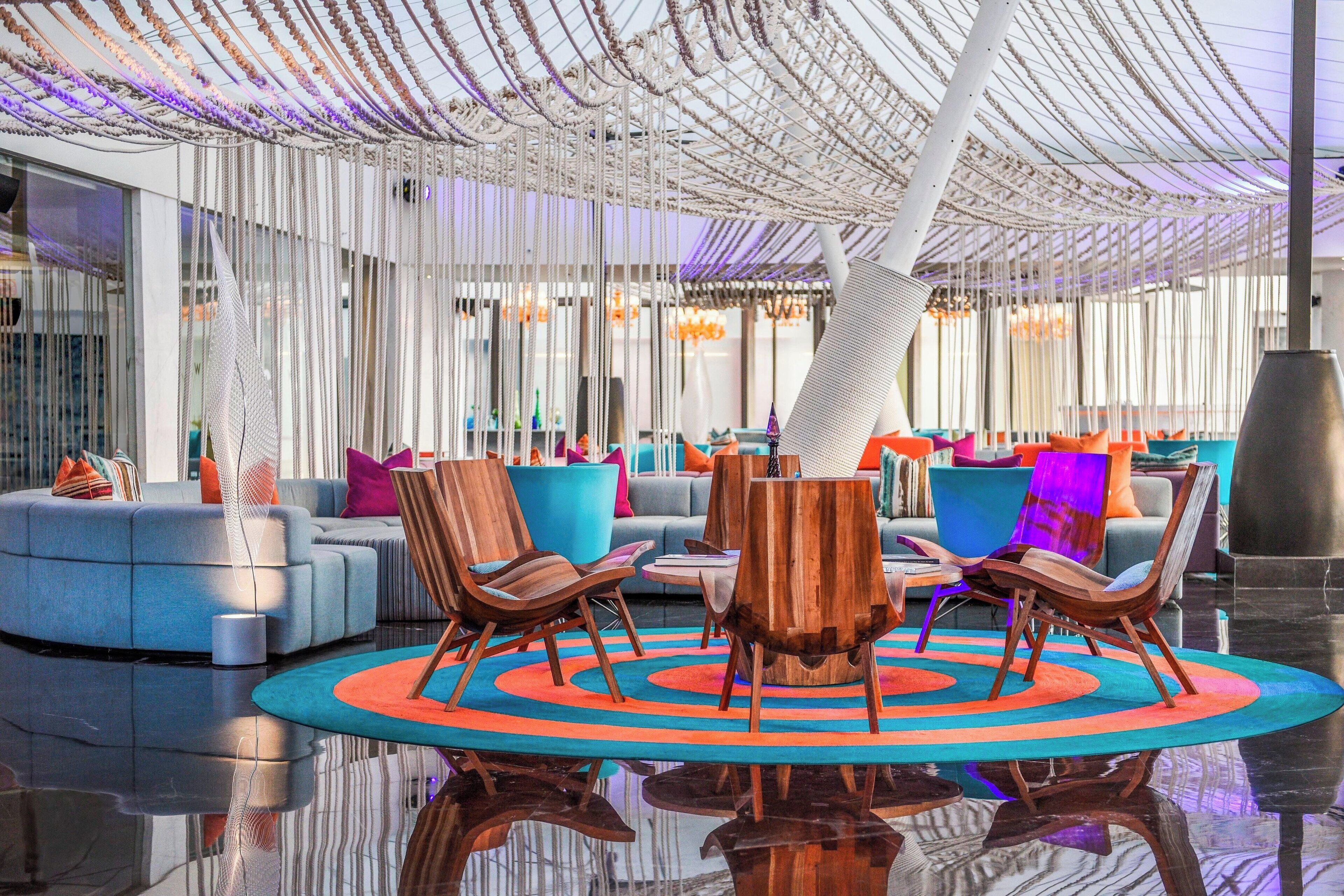 Un área de lounge moderna con piso reflectante, una mesa redonda central con sillas, y un espacio de descanso con sofás y cojines de colores.