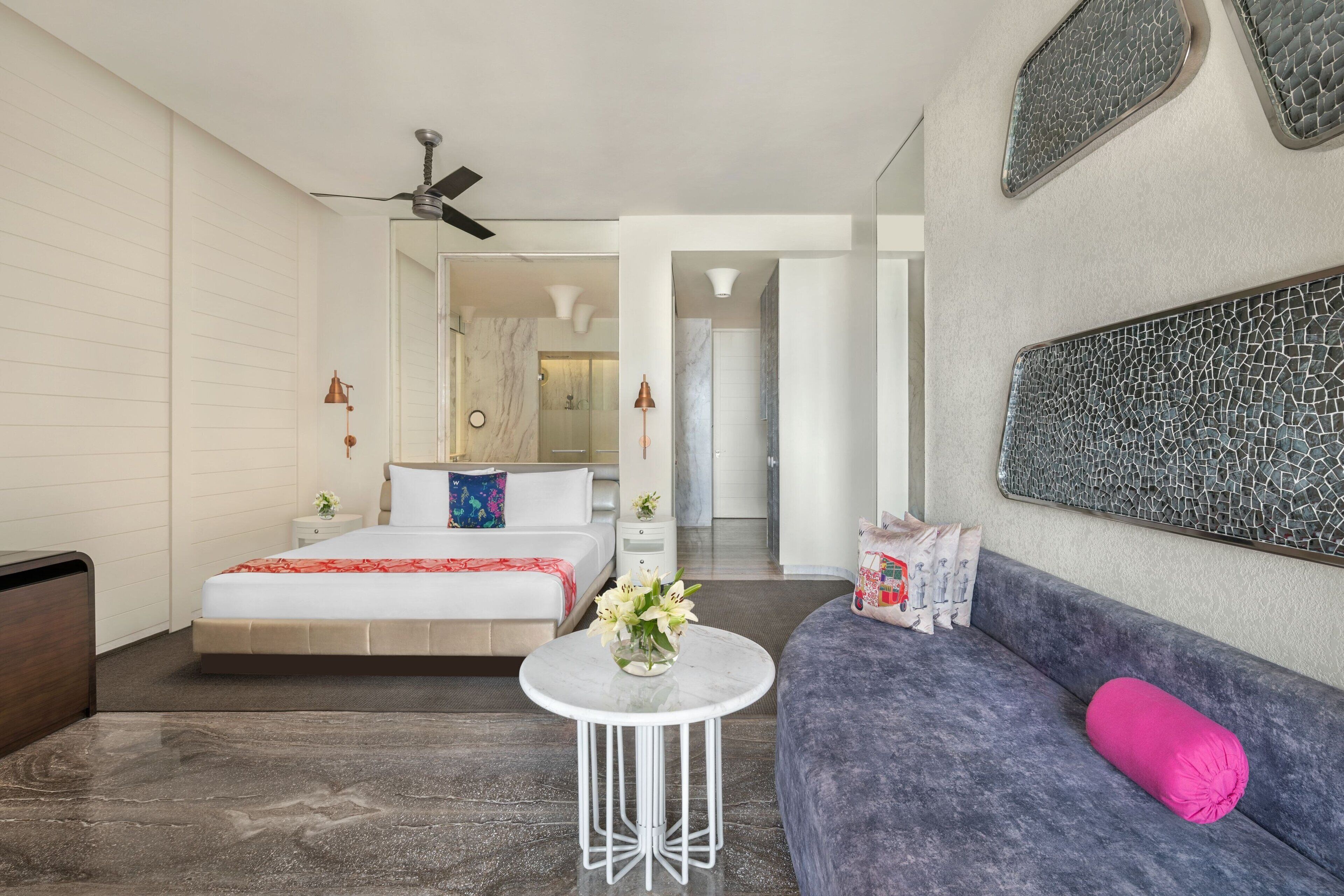 A modern hotel room with a bed, a sofa, a small table with a vase of flowers, and a ceiling fan.