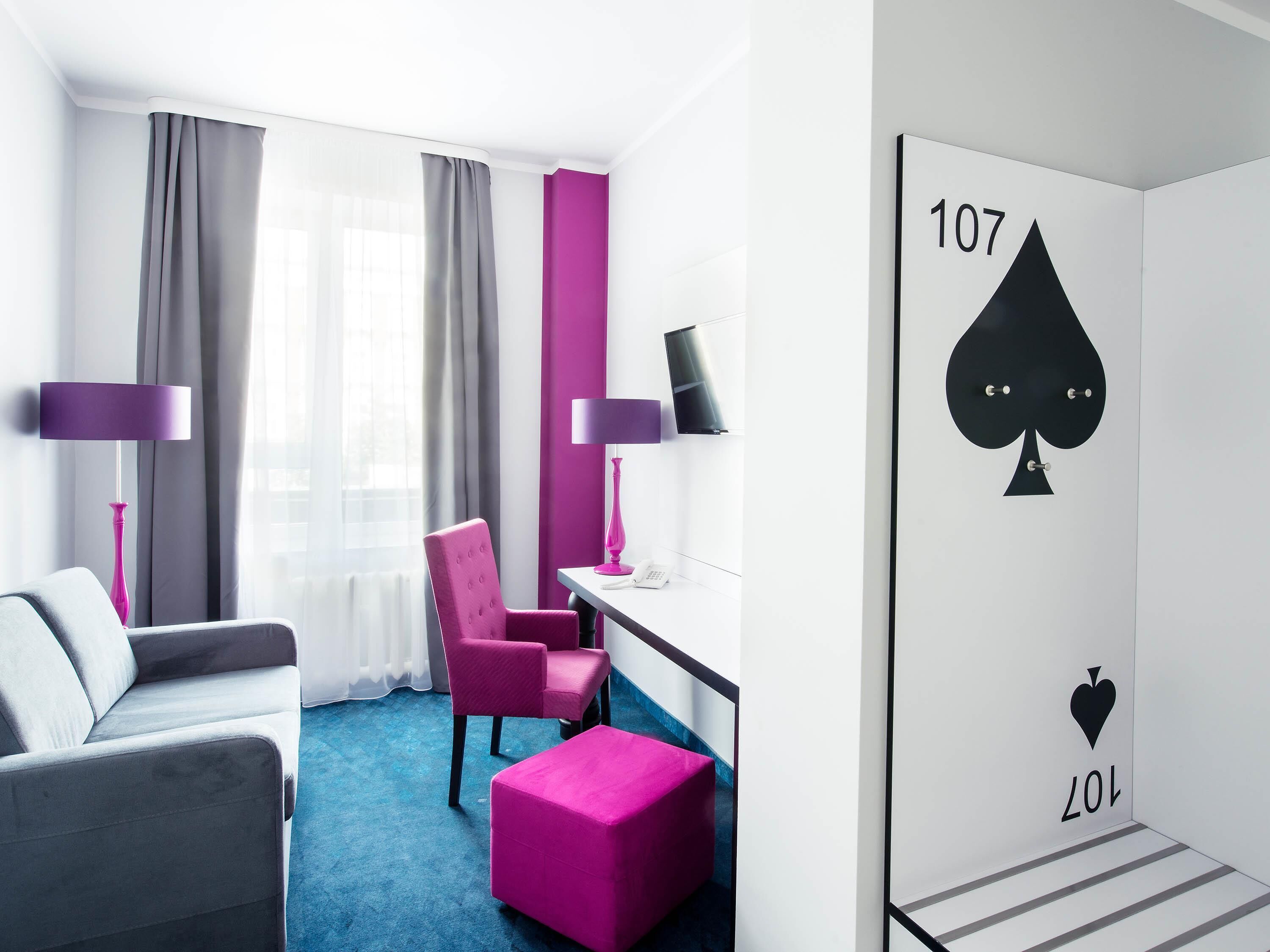 Habitación de hotel con sofá gris, una silla rosa, alfombra azul y una pared con un símbolo de pala.
