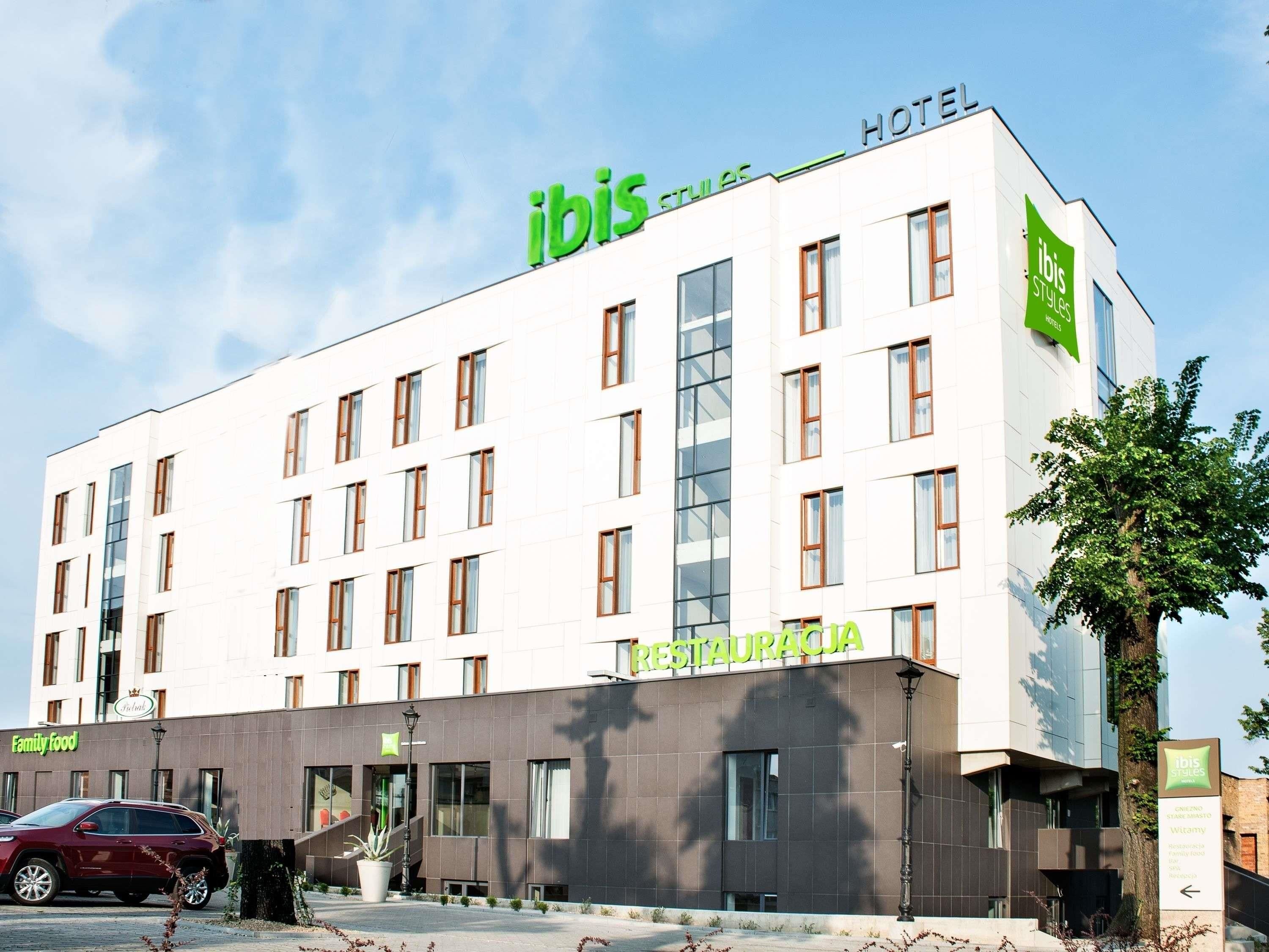 Ein modernes Hotelgebäude mit 'ibis Styles Hotels'-Beschilderung, ein Restaurant namens 'RESTAURACJA' im Erdgeschoss und ein Parkplatz mit einem roten Auto.