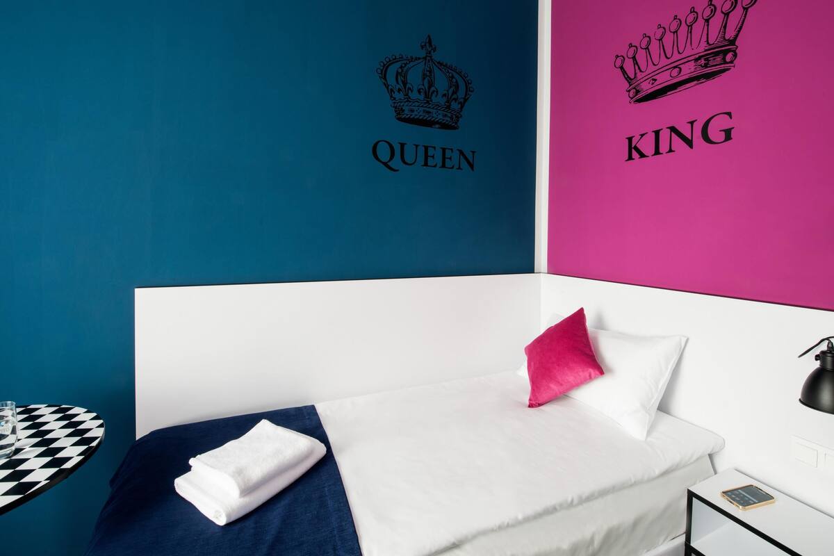 Ett sovrum med en säng, en kudde, ett nattduksbord, en lampa och en vägg med texten ”QUEEN” och ”KING”.