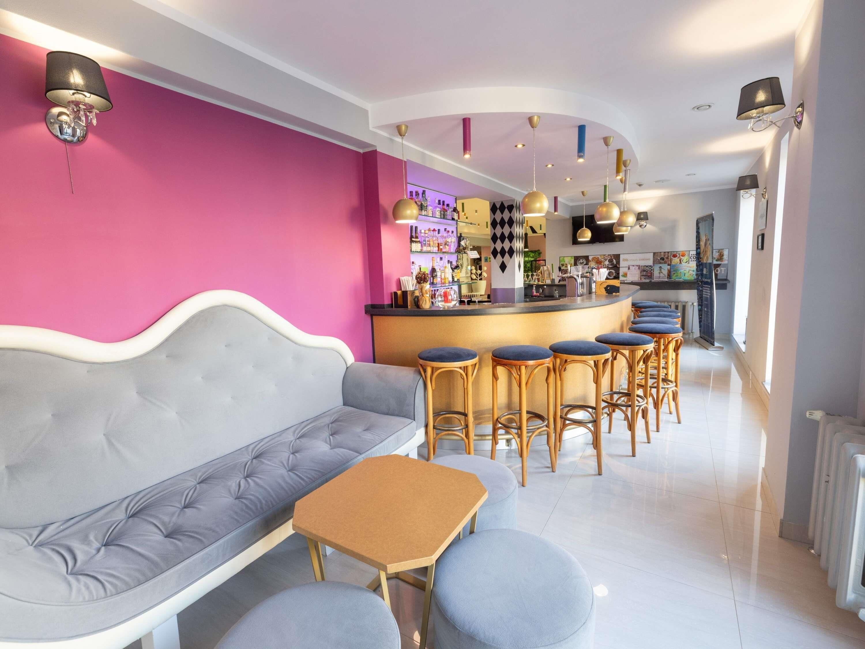 Uma área de bar moderna com parede rosa, sofá cinza e banquetas de madeira.