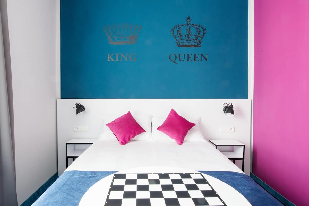 A bedroom with a blue and pink color scheme, featuring a bed with two pillows, a checkered blanket, and a wall with 'KING' and 'QUEEN' crowns.