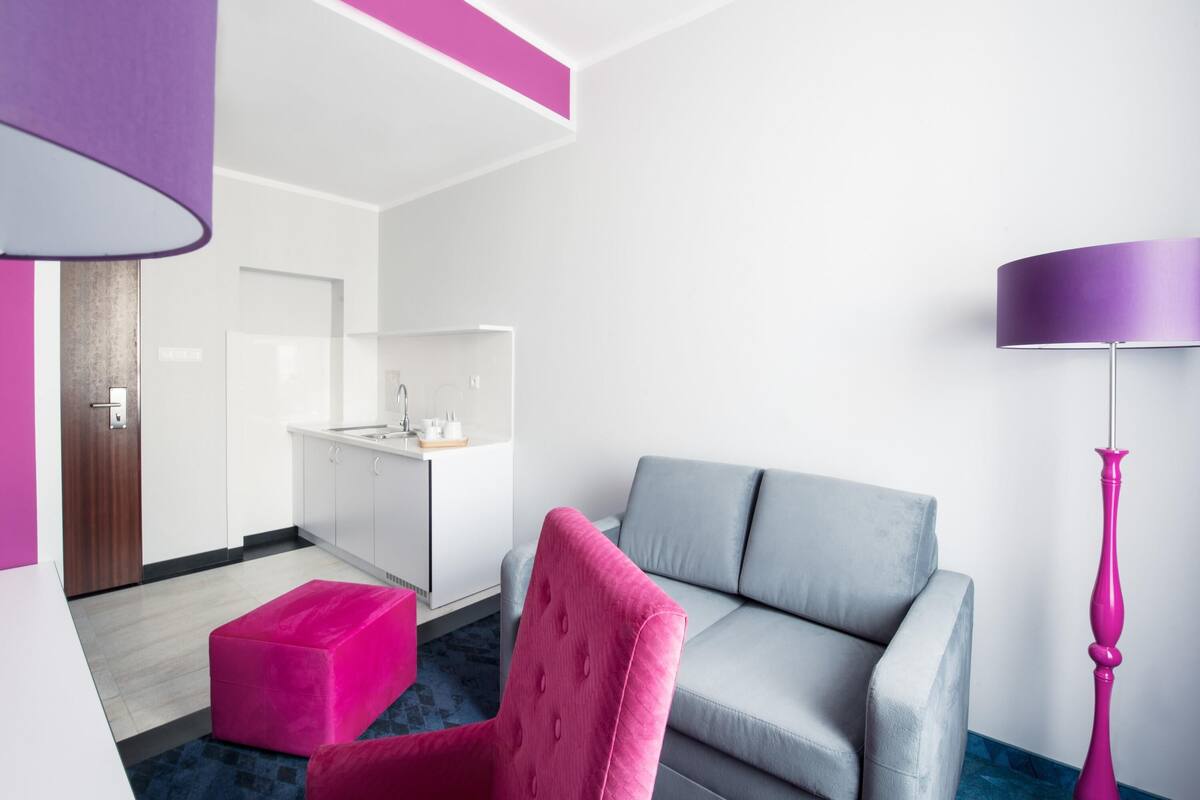 Ett modernt hotellrum med en grå soffa, en rosa fåtölj, en liten köksdel och en lila lampa.