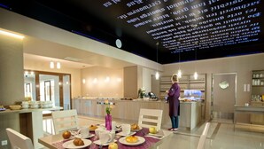 A modern kitchen with a dining area, a woman standing by the counter, and a digital menu display. - ibis Styles Gniezno Stare Miasto (Gniezno)