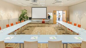 A conference room with long tables, orange chairs, a projector screen, and a plant. - ibis Styles Gniezno Stare Miasto (Gniezno)