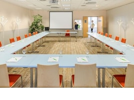 Uma sala de conferência com mesas compridas, cadeiras laranja, uma tela de projetor e uma planta.