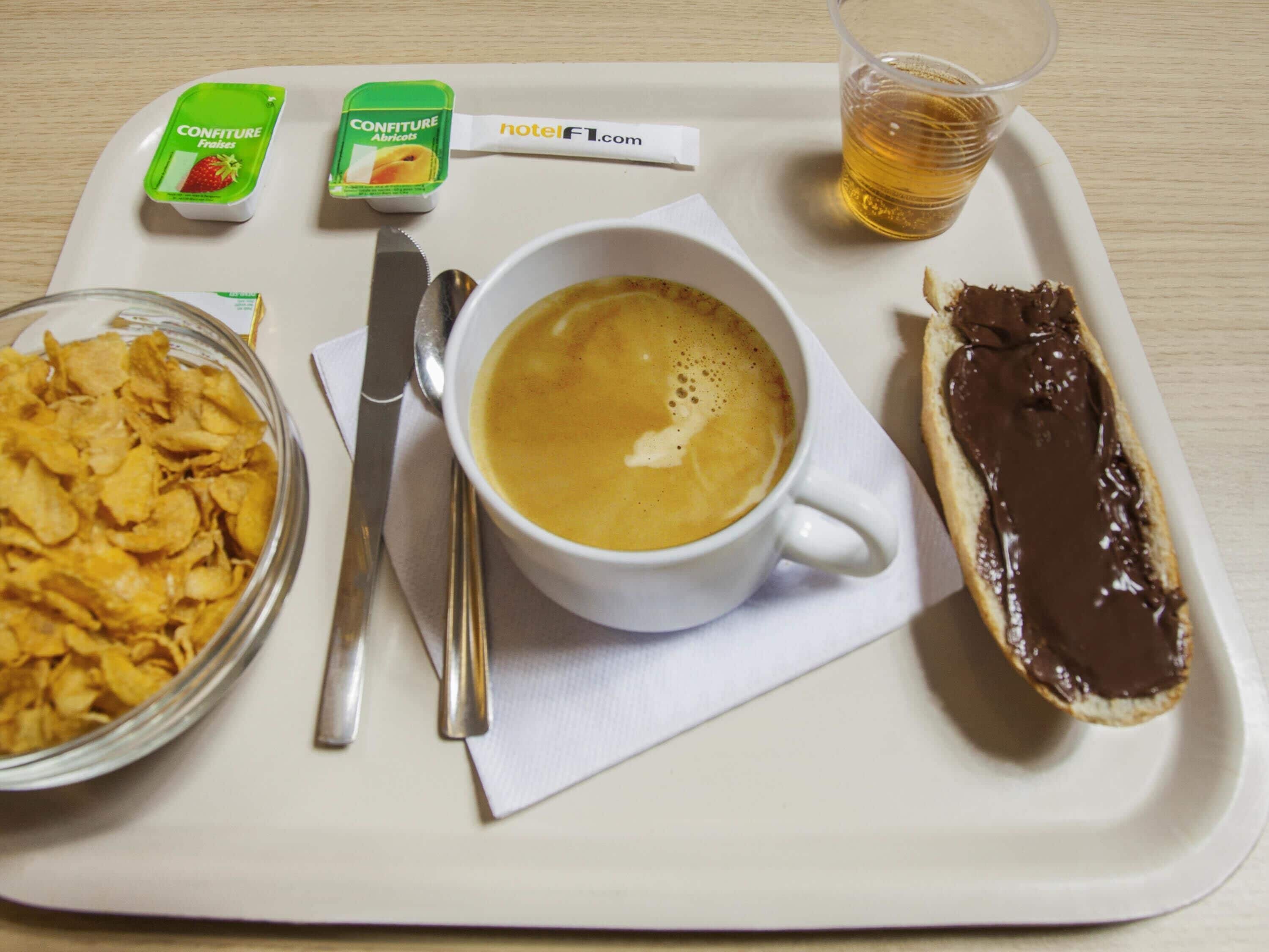 Una bandeja con una taza de café, un tazón de cereal, un pastelito cubierto de chocolate y dos recipientes de yogur.