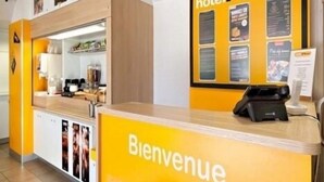 Zona de recepción de hotel con un cartel que dice 'Bienvenue', una mesa de recepción, un teléfono y carteles informativos en la pared.