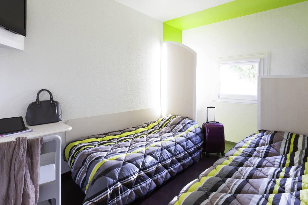 A hotel room with two beds, a desk, a chair, a suitcase, and a window.