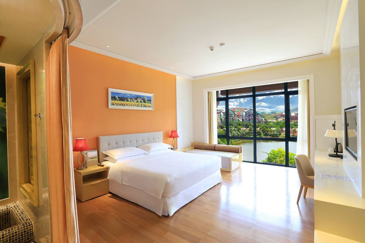 A modern hotel room with a large bed, a desk, and a view of a cityscape through large windows.