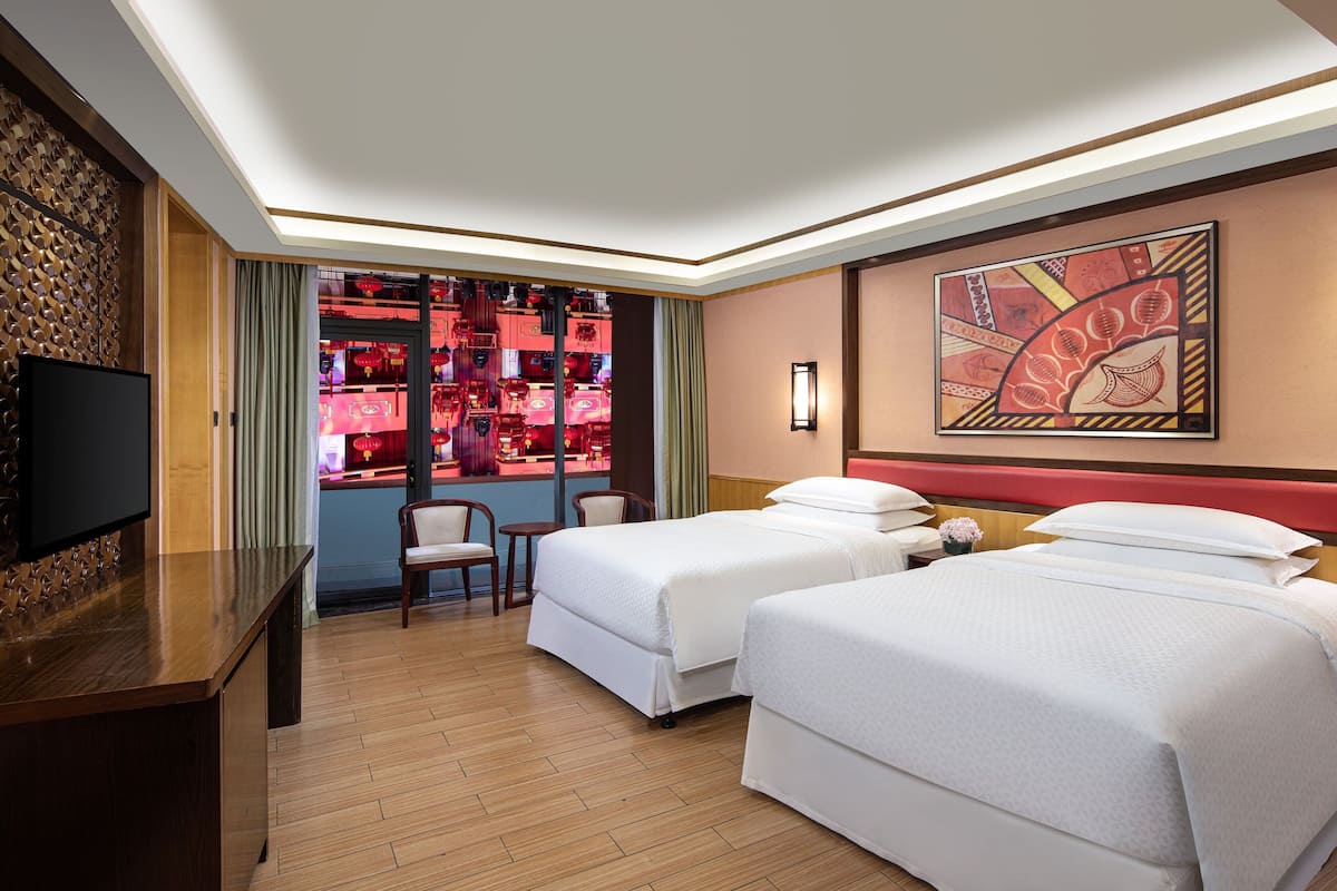 A hotel room with two beds, a TV, a desk, and a view of a restaurant with red decor.