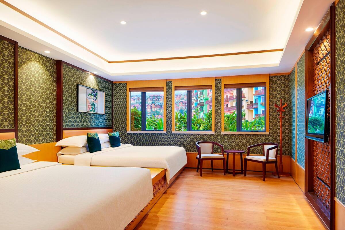 A hotel room with two beds, a desk, chairs, large windows with a view, and a TV.