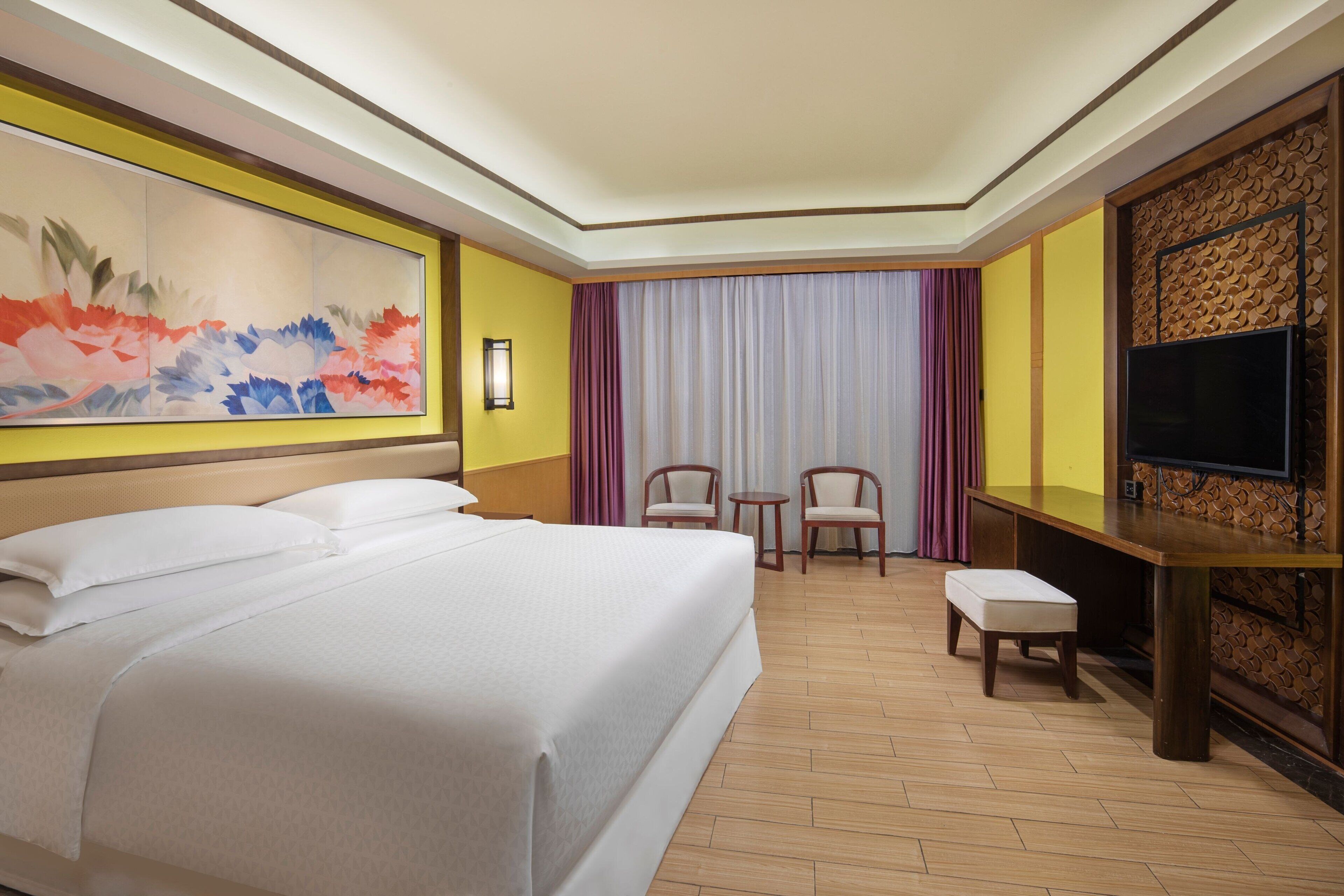 A hotel room with a large bed, two chairs, a small table, a TV, and a decorative wall panel.