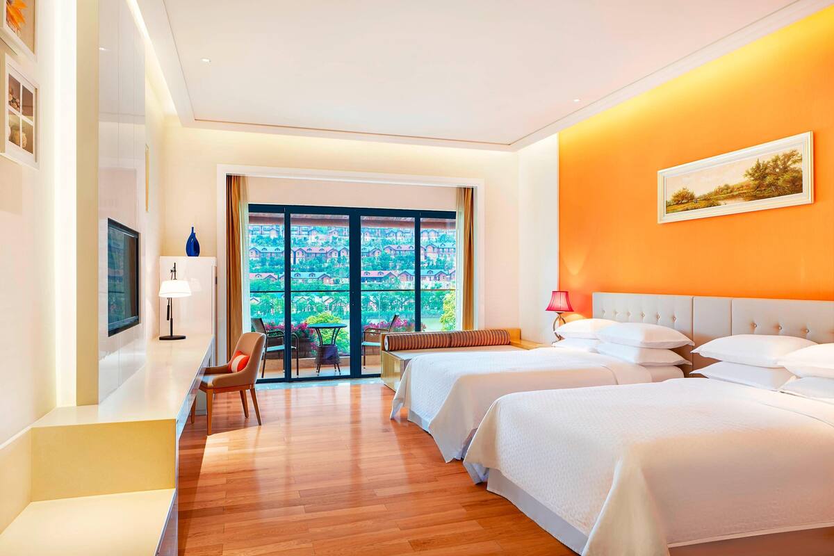 A hotel room with two beds, a desk, a chair, and a large window with a view of greenery.