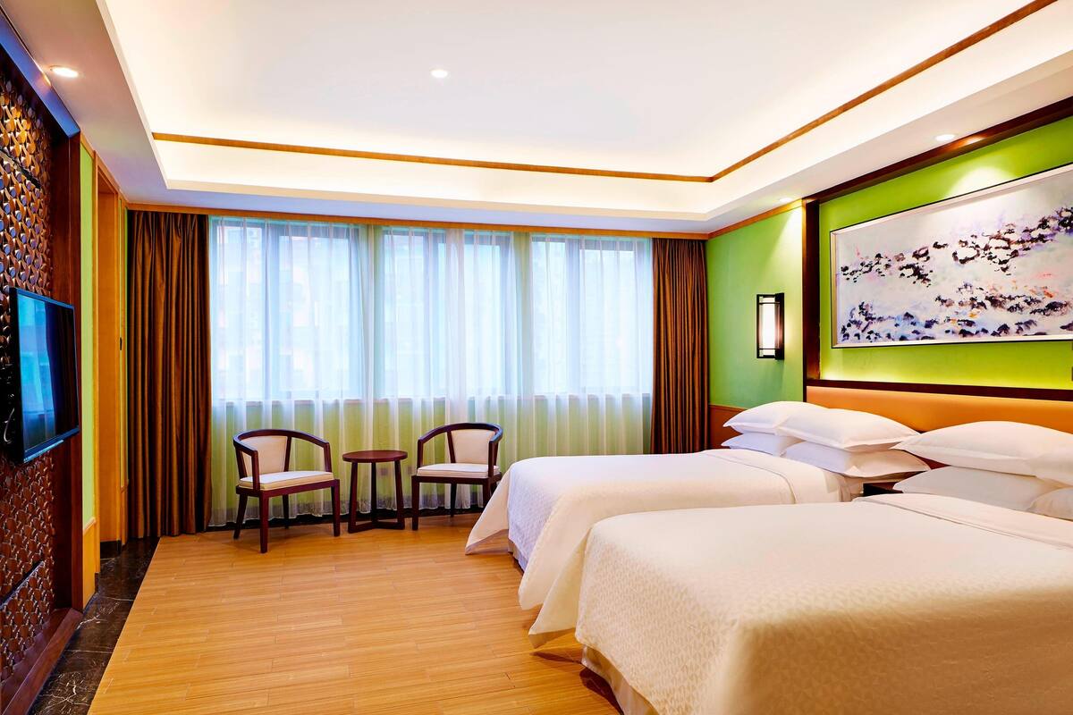A hotel room with two beds, a TV, a painting, and a view of the outside through sheer curtains.
