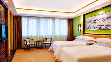 A hotel room with two beds, a TV, a painting, and a view of the outside through sheer curtains.