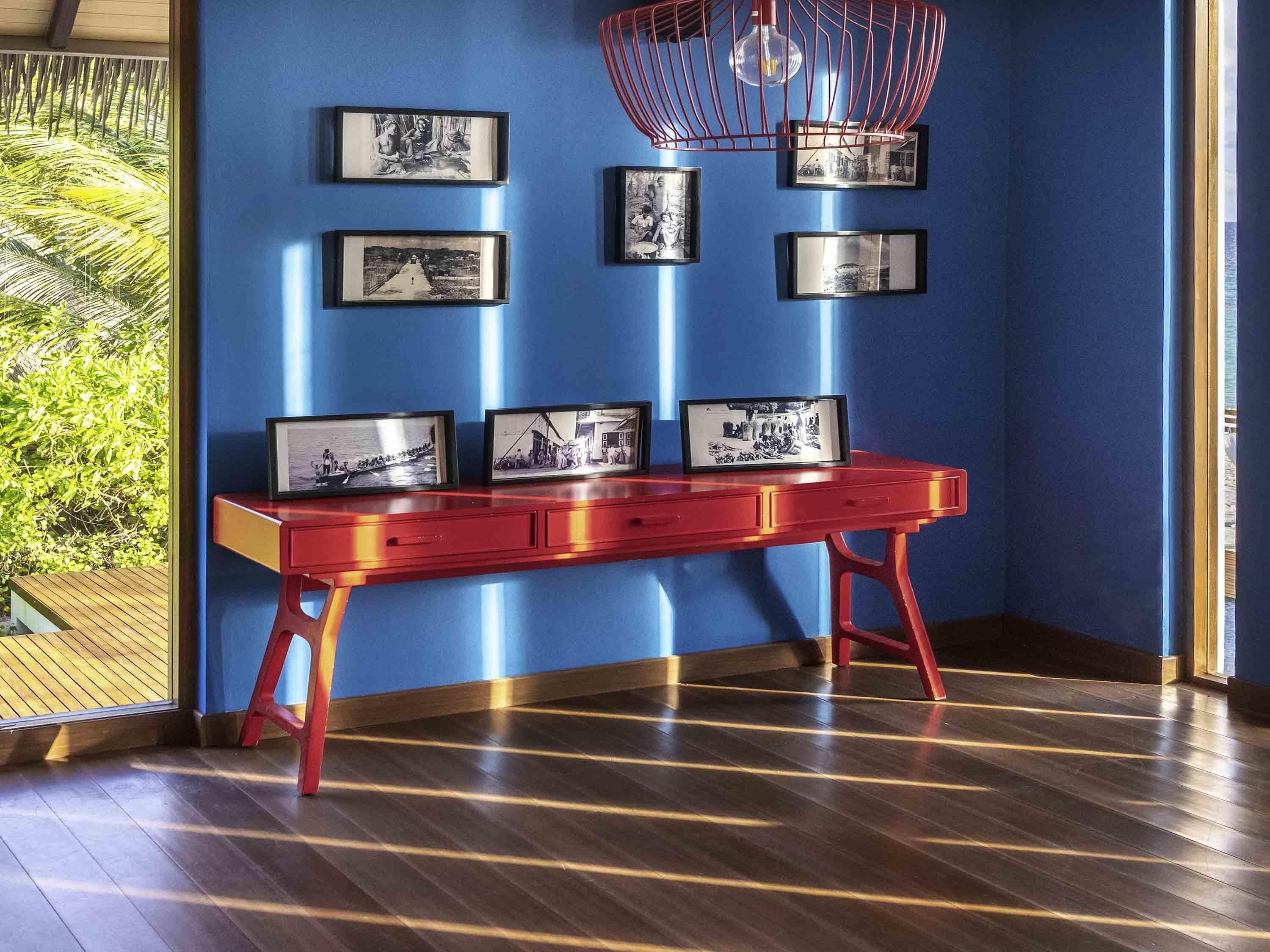 Una stanza con pavimento in legno, una scrivania rossa con cassetti e un muro blu con quadri appesi.