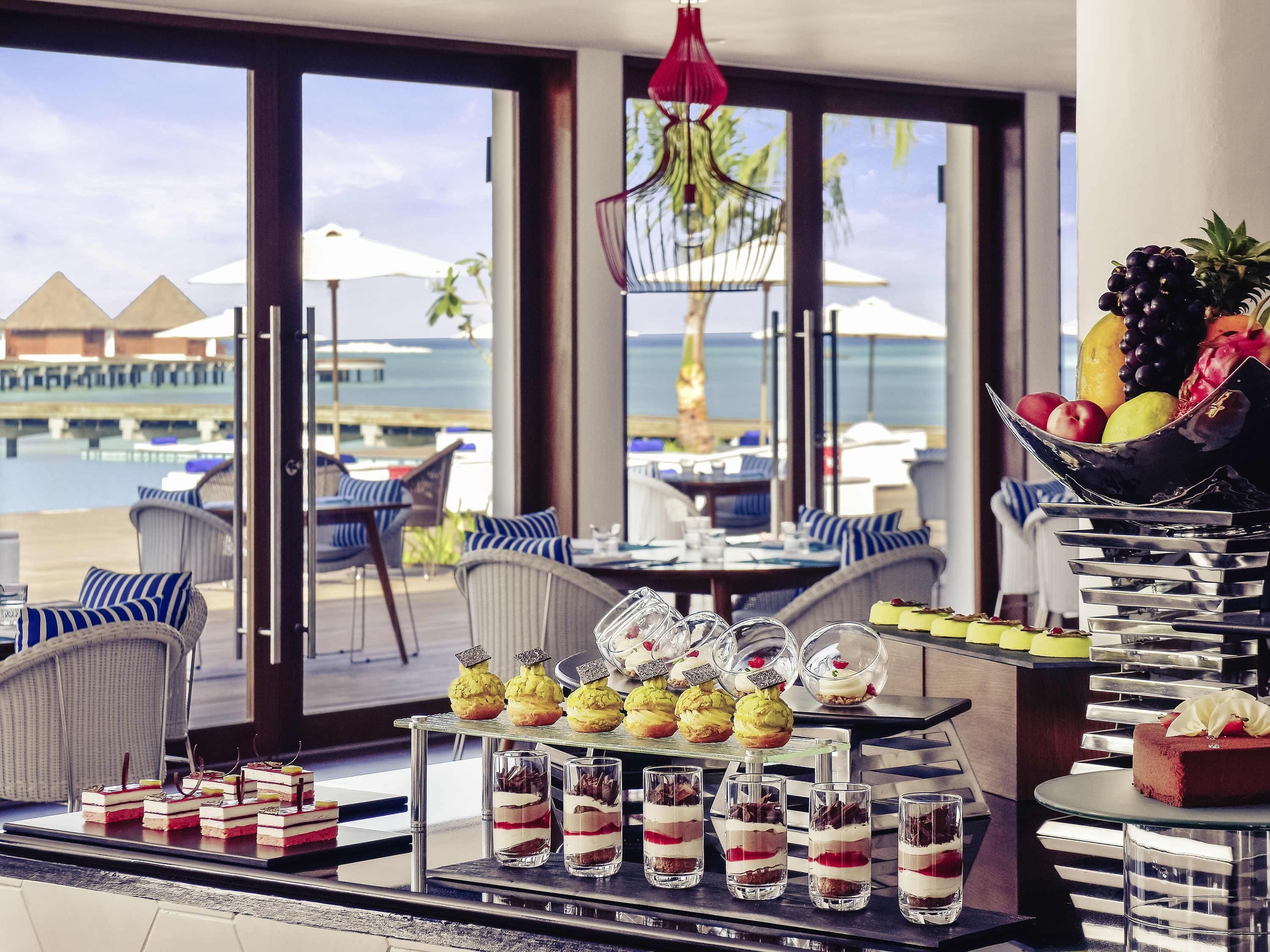 Una mesa de buffet con postres y un bol de frutas, con vista a una playa con sombrillas y sillas.