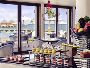 A buffet table with desserts and a fruit bowl, overlooking a beach with umbrellas and chairs.