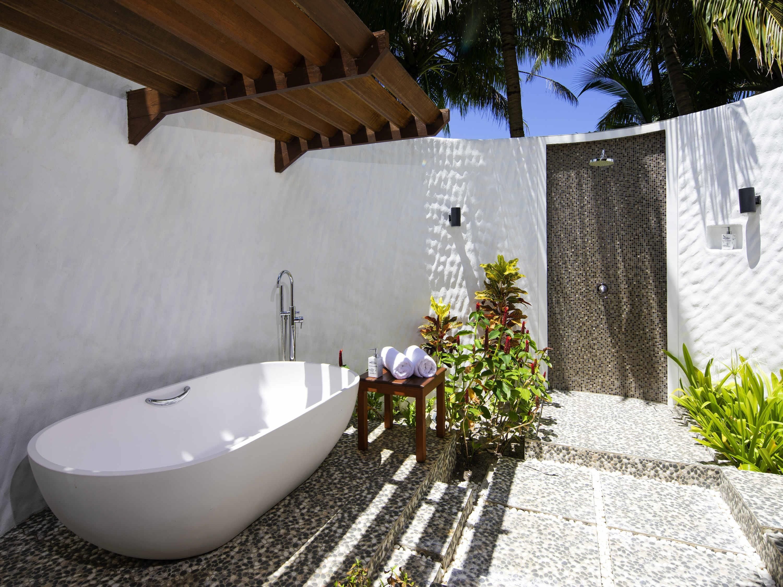 A modern outdoor bathroom with a freestanding bathtub, a small wooden stool with towels, and a mosaic wall.