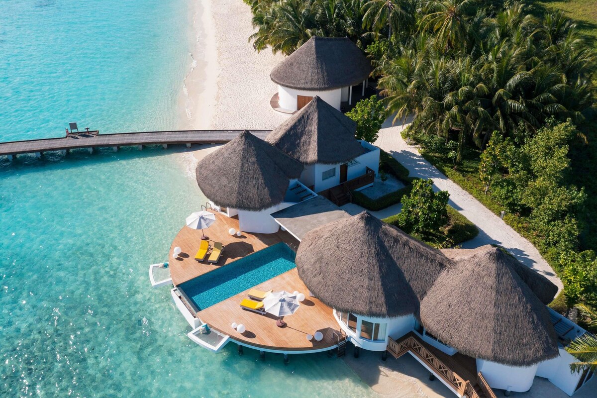 An overwater bungalow with a pool, thatched-roof structures, and a beach view.