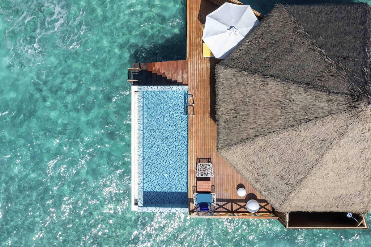 Vue aérienne de bungalows sur pilotis dotés d’une piscine, de chaises longues et d’un parasol en feuilles de palmier.