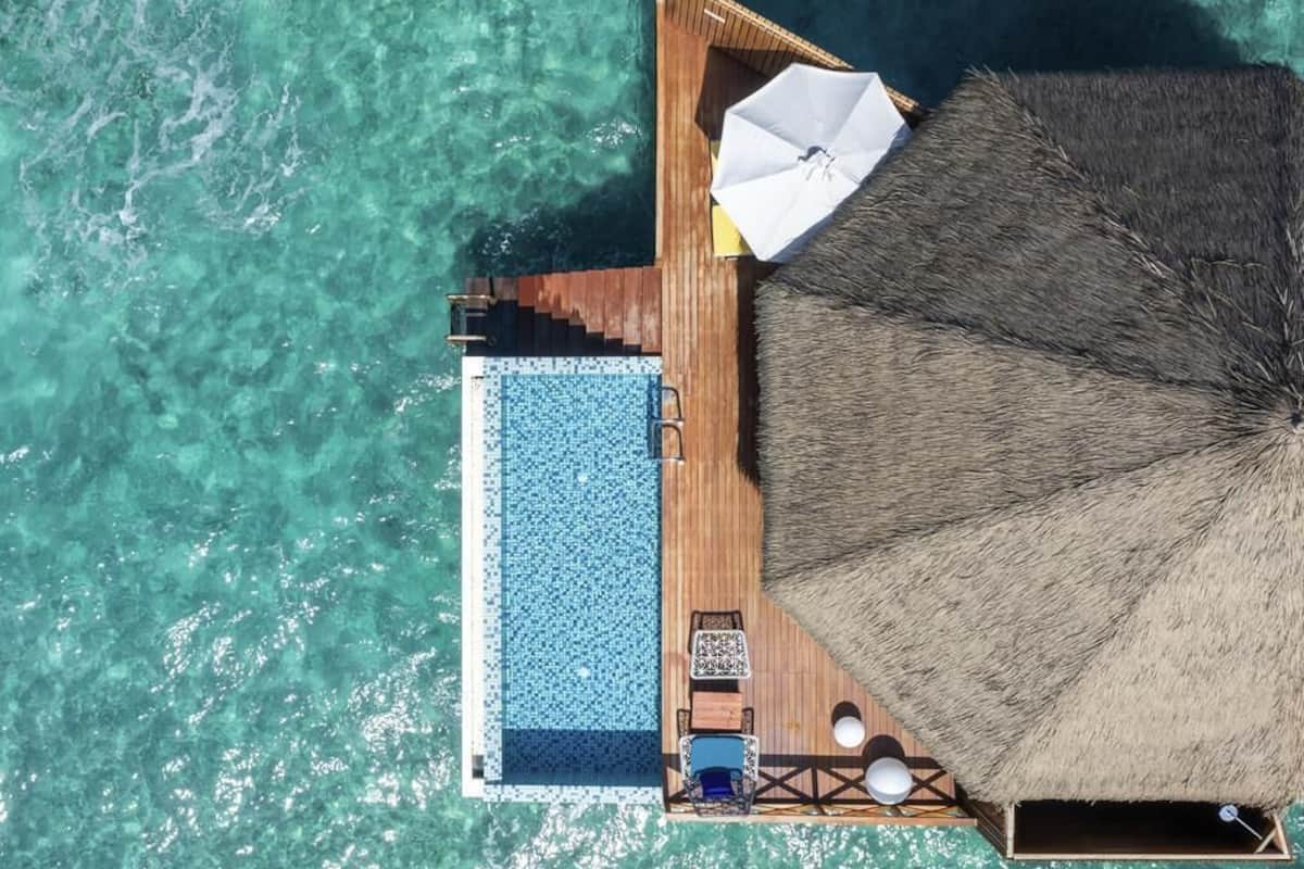 An aerial view of an overwater villa with a swimming pool, lounge area, and thatched-roof structure.