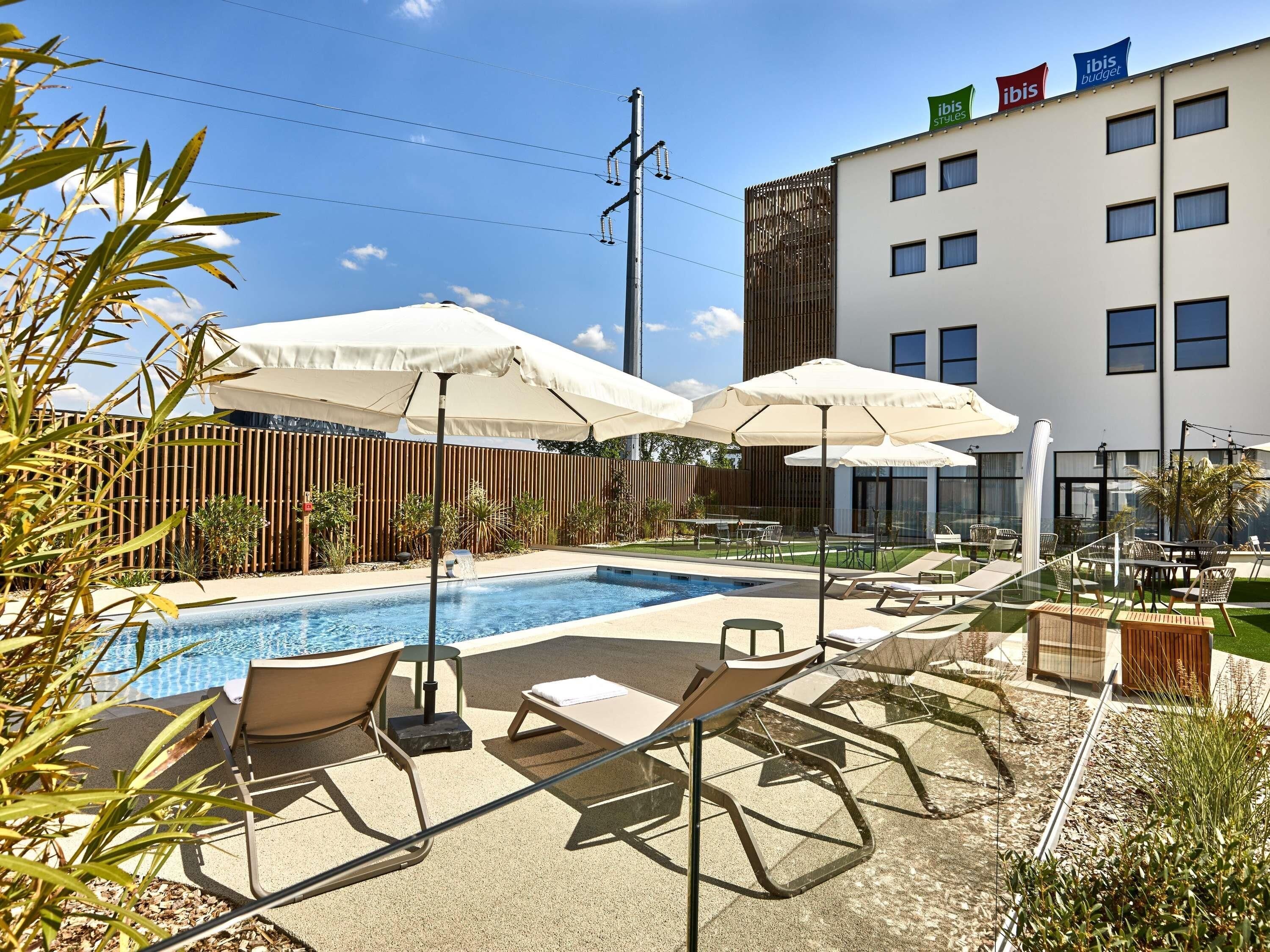 A pool area with lounge chairs and umbrellas, a wooden fence, and a modern building with flags.
