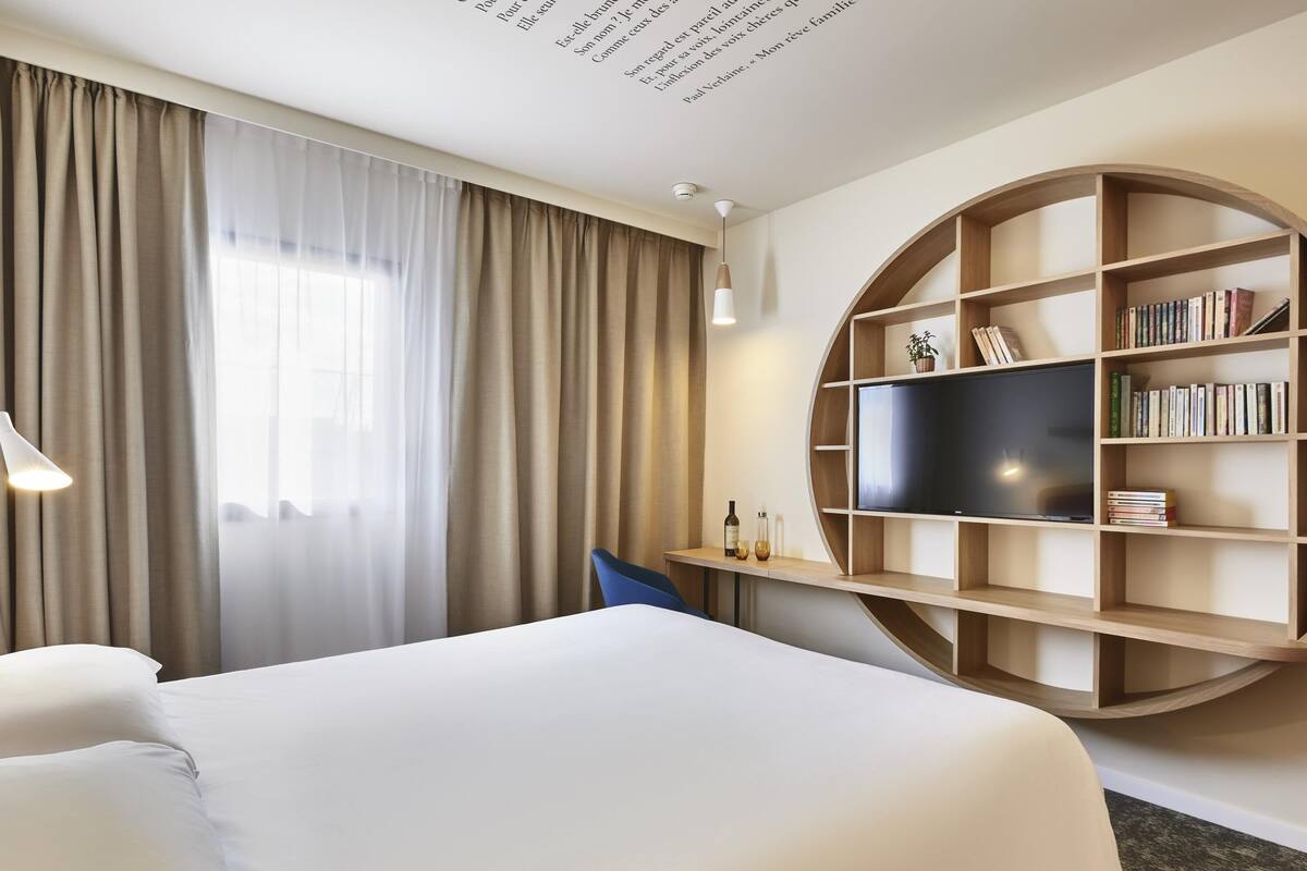 A hotel room with a bed, a television, a bookshelf, and a desk with a lamp.