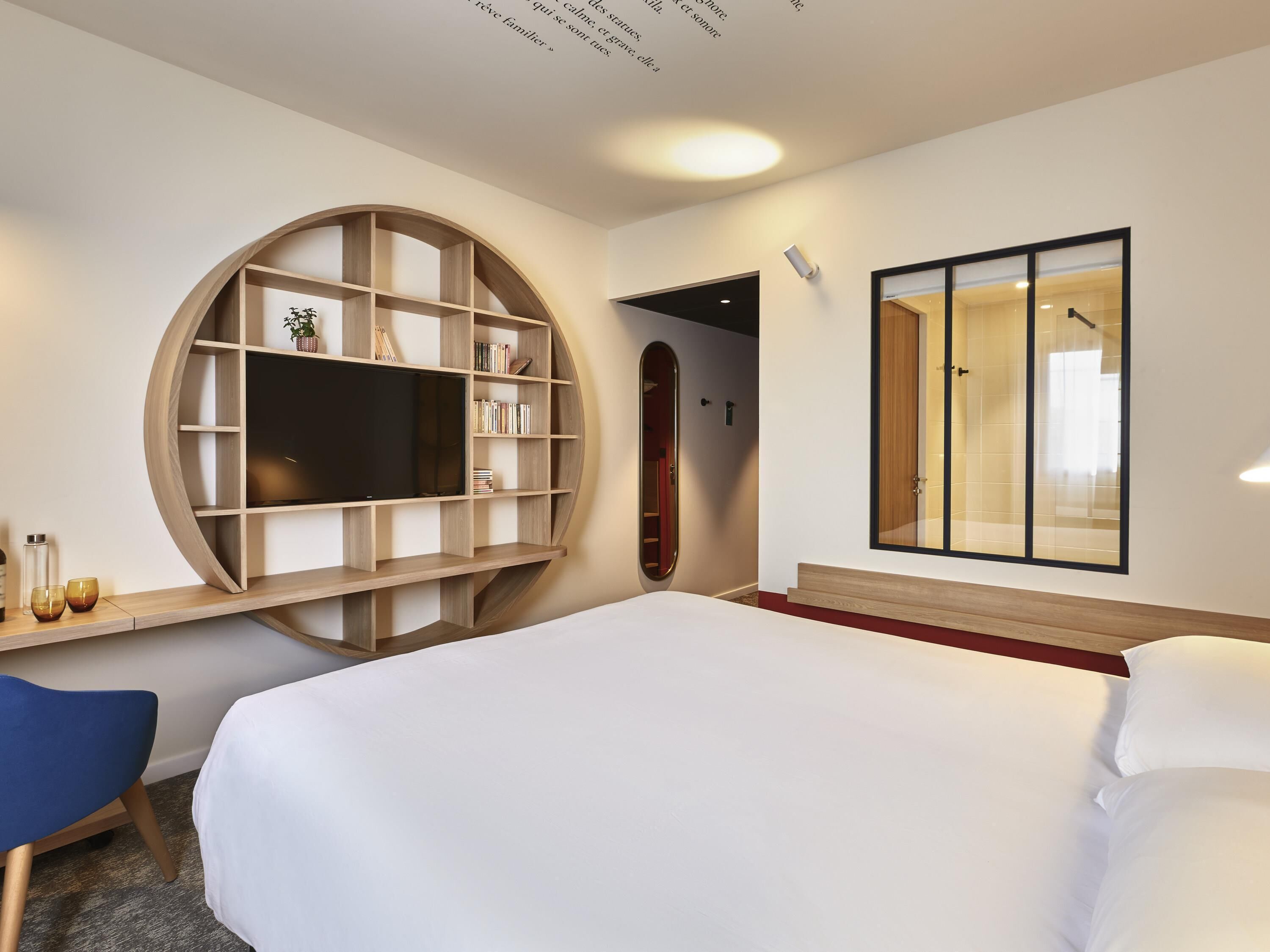 A hotel room with a large bed, a TV mounted on a circular wooden shelf, a wardrobe with a mirror, and a small desk with a chair.