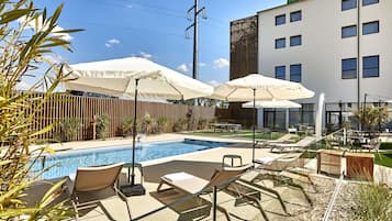 A pool area with lounge chairs and umbrellas, a wooden fence, and a modern building with flags.