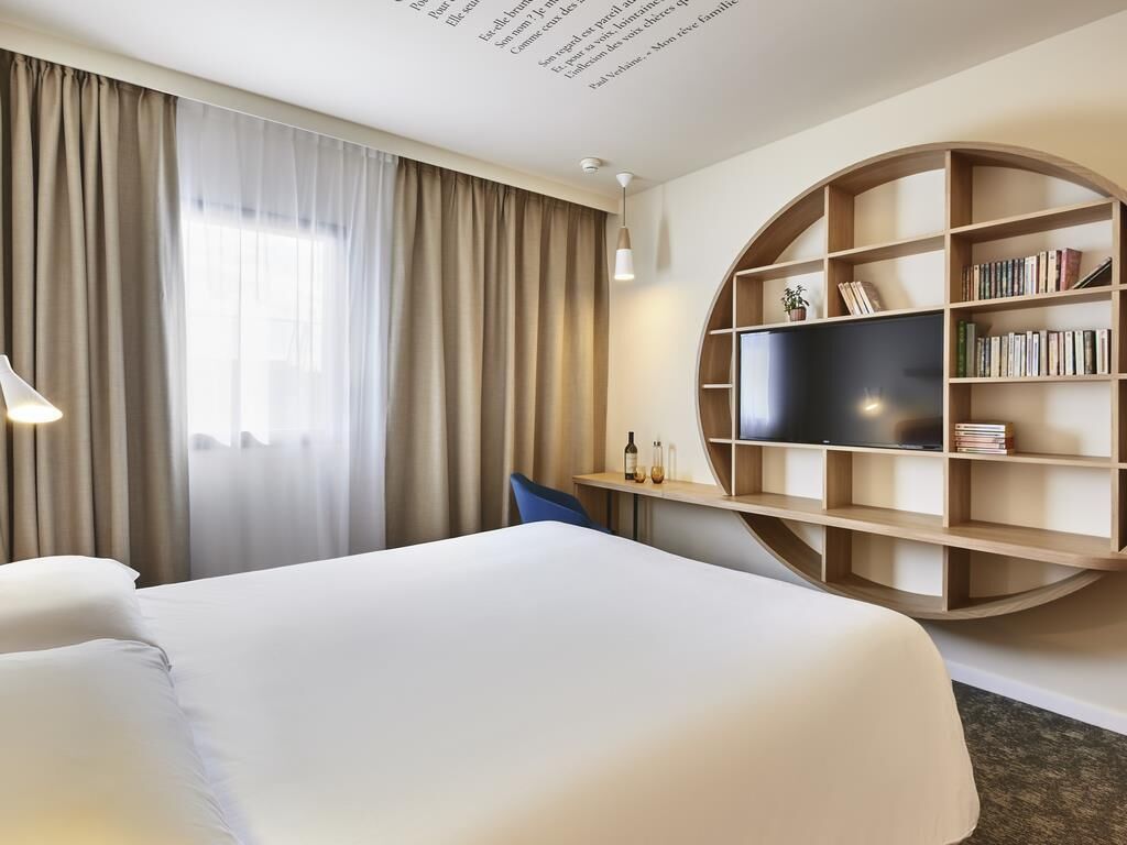 A hotel room with a bed, a TV, a bookshelf, and a window with curtains.