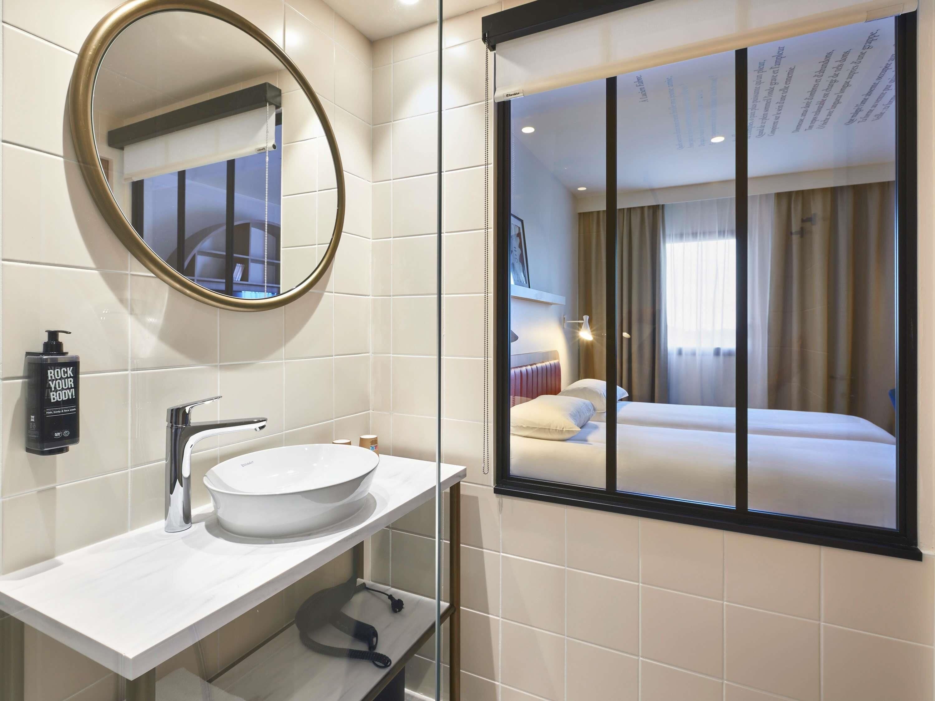 Bany d'hotel amb un mirall rodó, un lavabo blanc i una finestra amb vista a un dormitori.