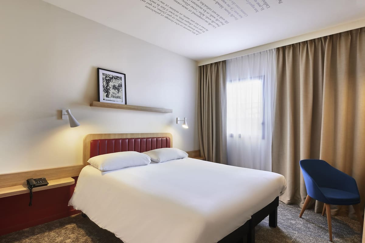 A hotel room with a bed, a desk with a telephone, a chair, a wall-mounted shelf with a framed picture, and a window with curtains.