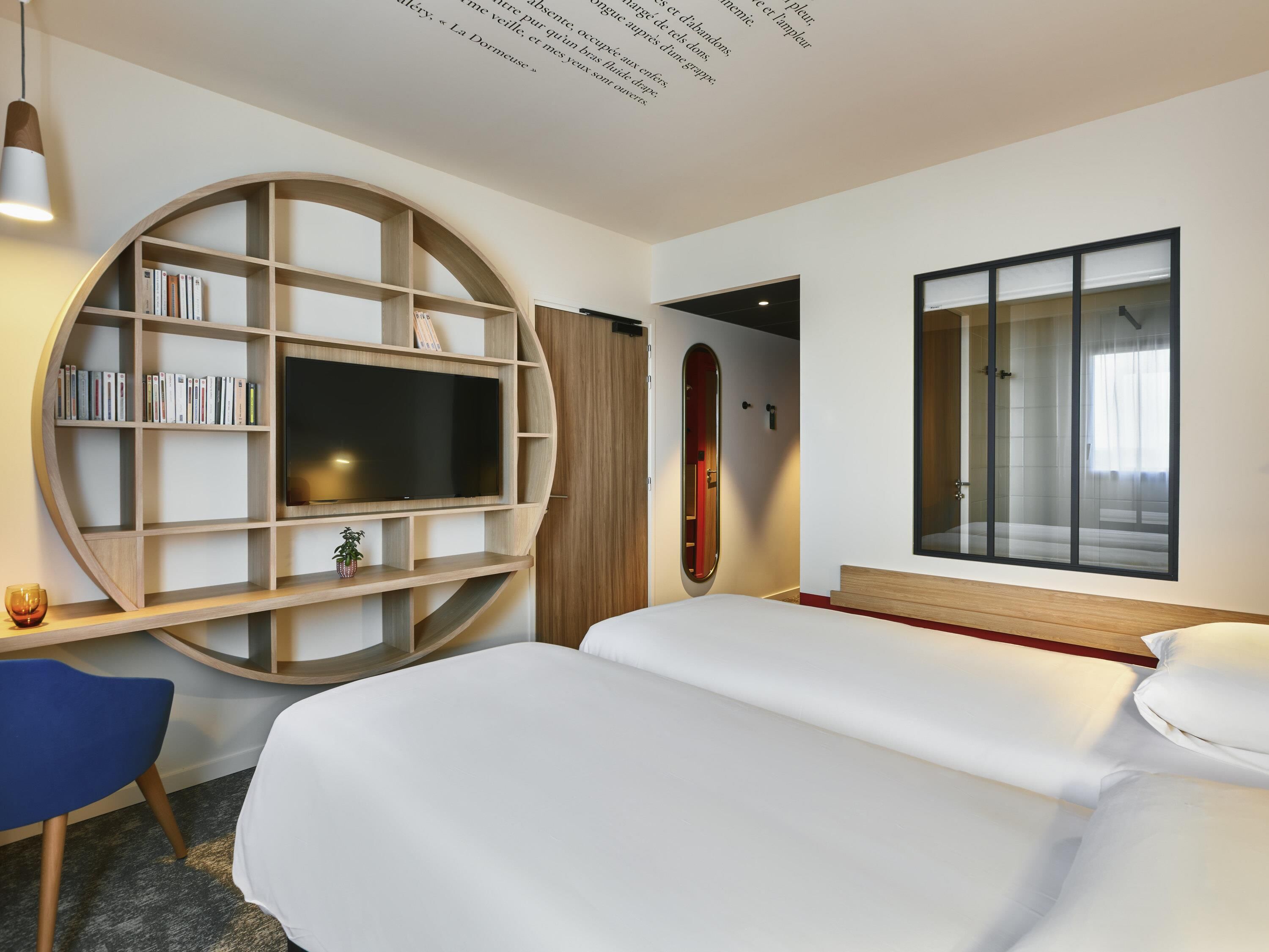 A modern hotel room with a large bed, a TV mounted on a curved wooden shelf, a small desk with a chair, and a large window with a view.
