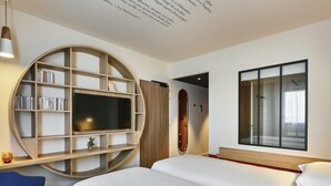 A modern hotel room with a large bed, a TV mounted on a curved wooden shelf, a small desk with a chair, and a large window with a view. - ibis Styles La Roche-sur-Yon (Mouilleron-le-Captif)