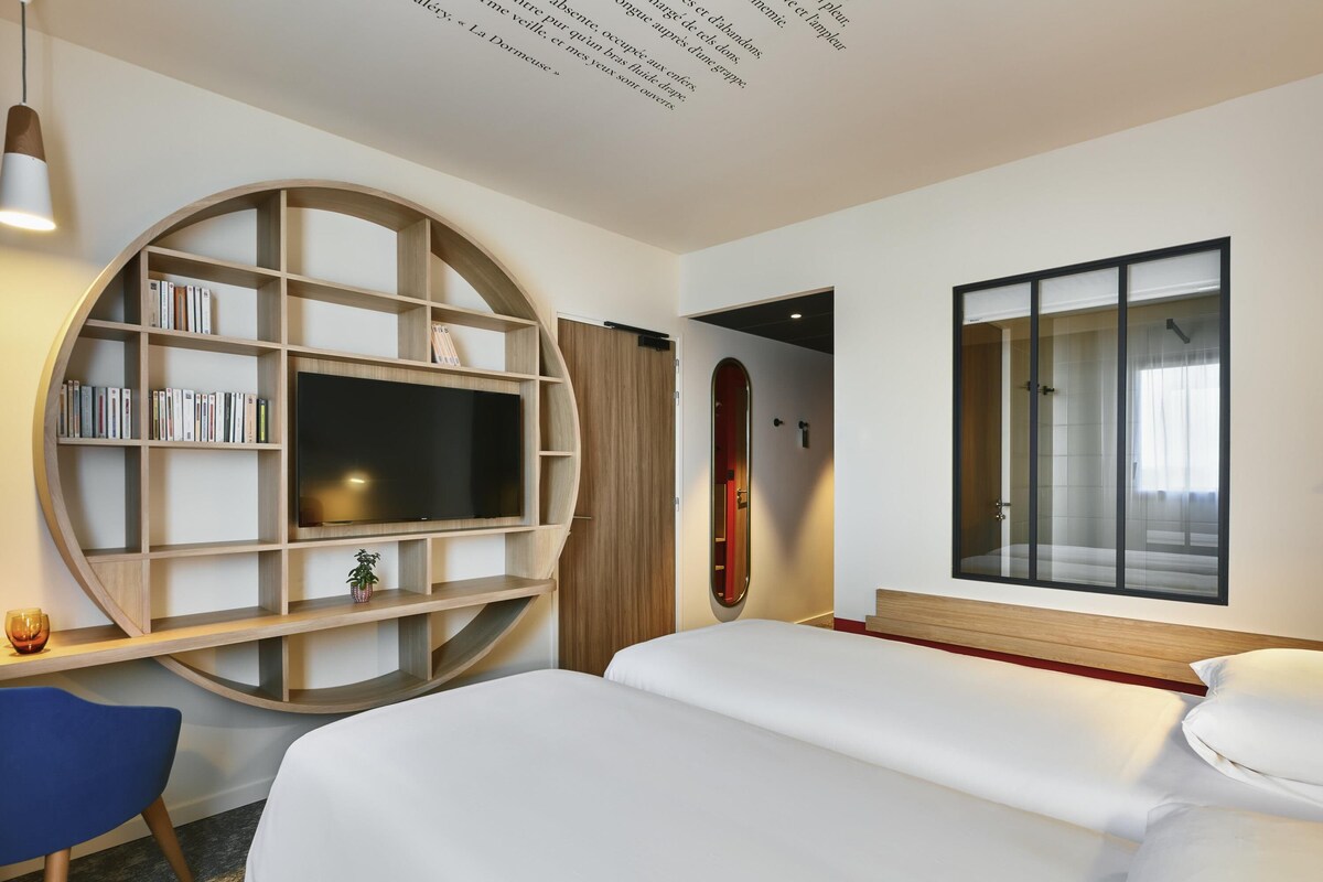 A modern hotel room with a large bed, a TV mounted on a curved wooden shelf, a small desk with a chair, and a large window with a view.