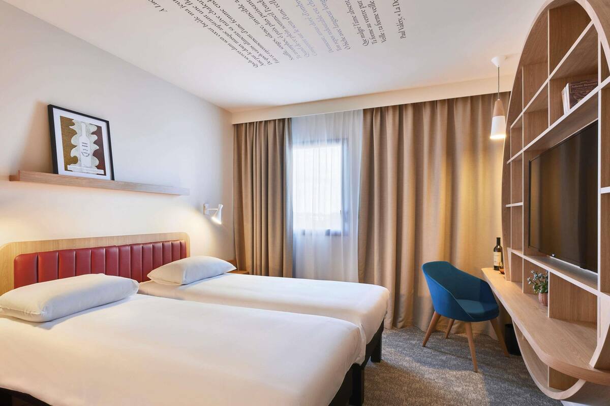 A hotel room with two beds, a desk, a chair, a TV, and a bookshelf.