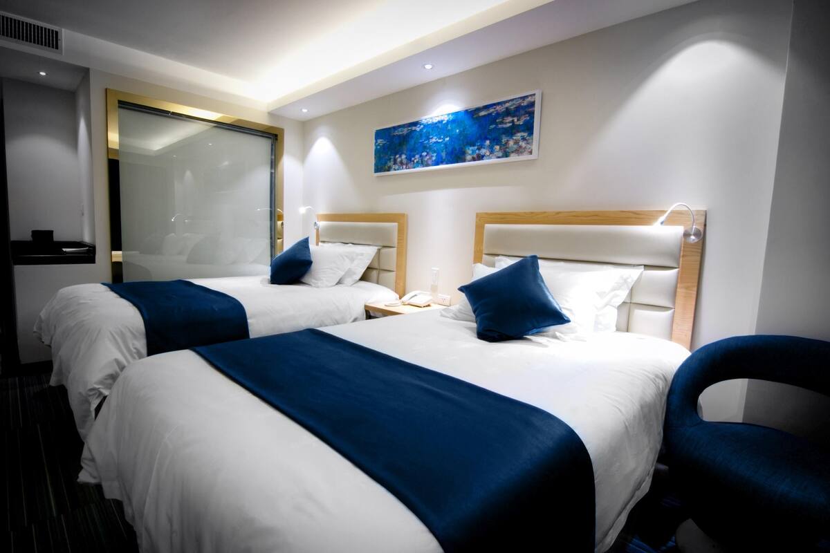A hotel room with two beds, a large mirror, a painting on the wall, and a desk with a lamp.