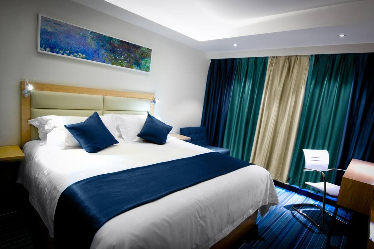 A hotel room with a large bed, a desk, a chair, a painting on the wall, and a view of the outside through curtains.