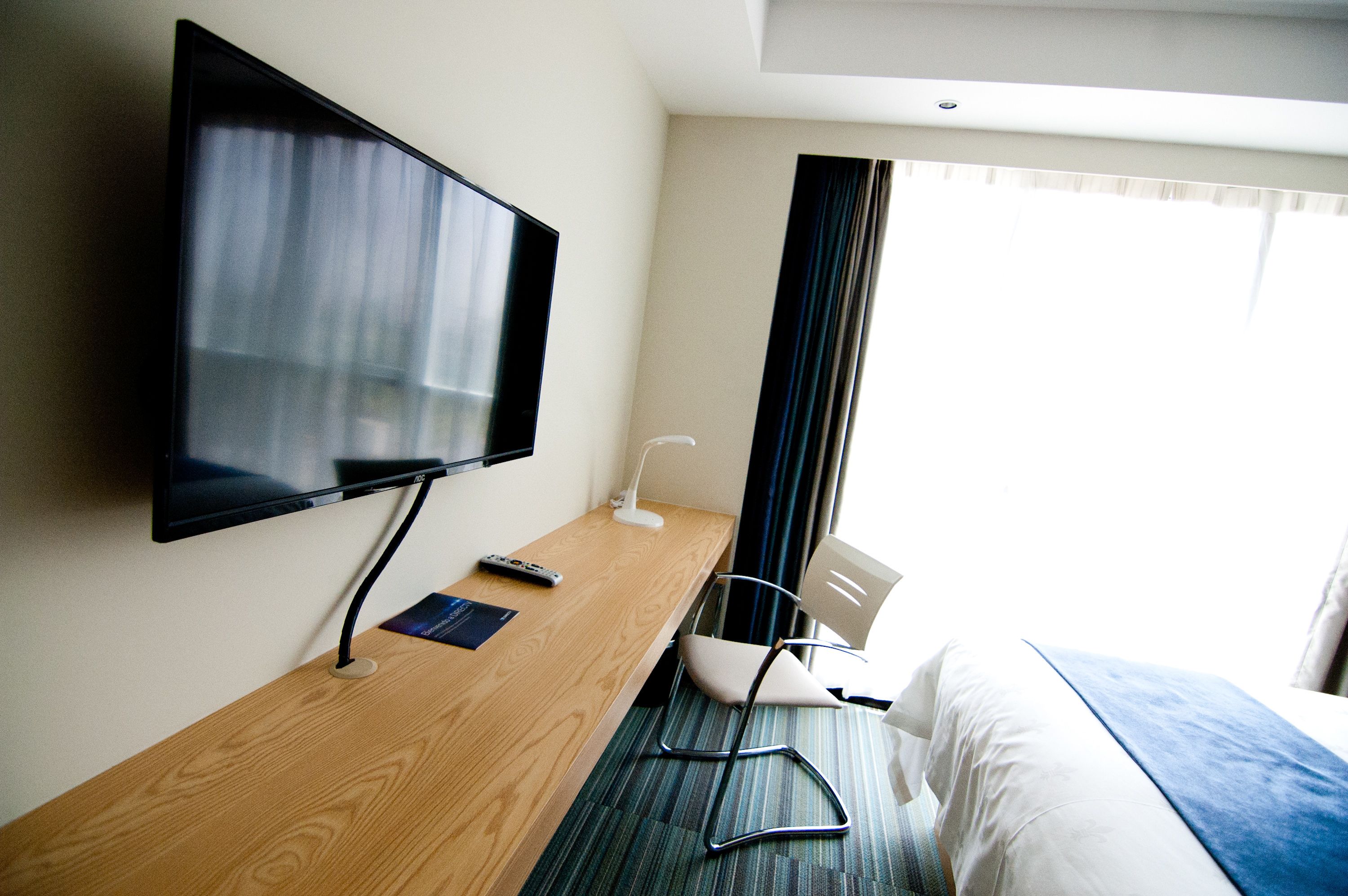 Habitación de hotel con televisión de pantalla plana en la pared, un escritorio de madera con un control remoto, una silla y una cama con manta azul.
