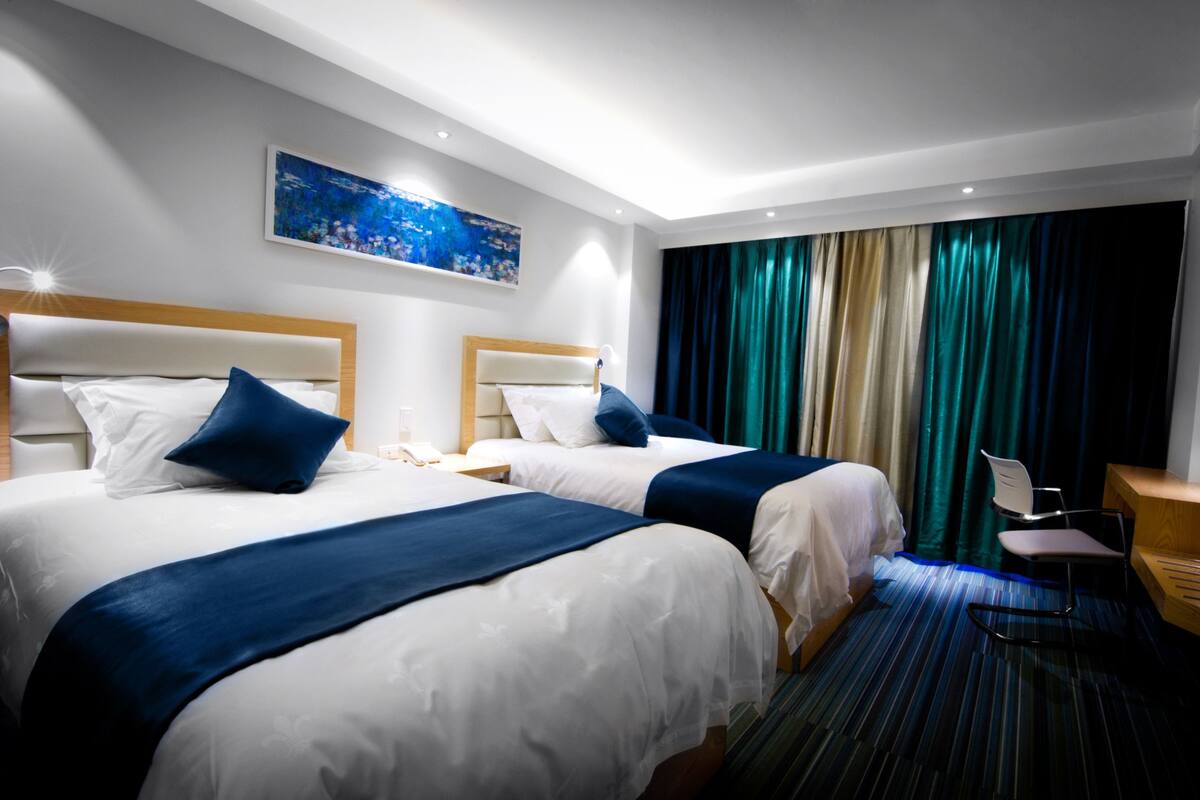A hotel room with two beds, a desk, and a chair. There is a painting on the wall and a window with curtains.