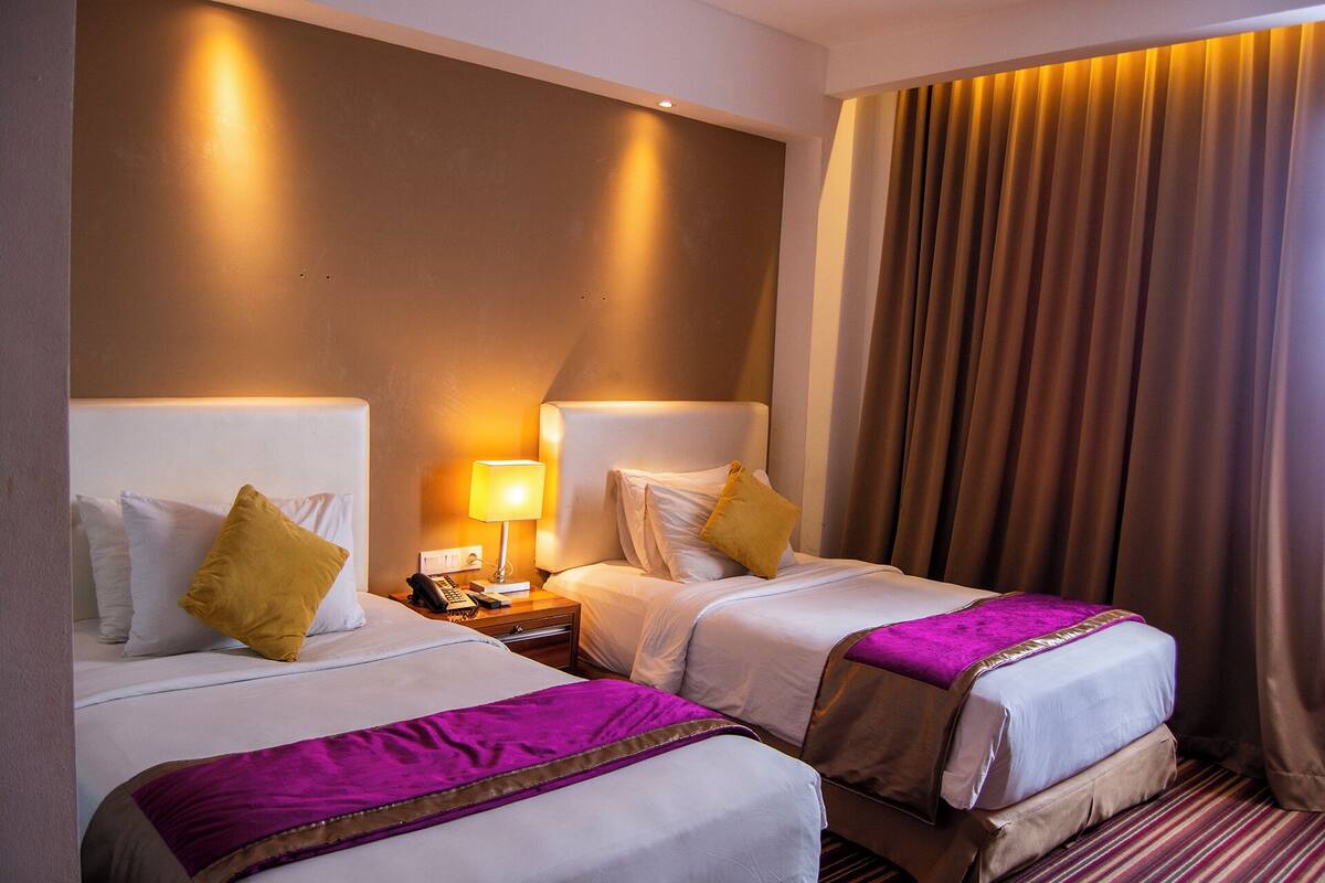 A hotel room with two beds, a nightstand with a lamp, and a large window with curtains.